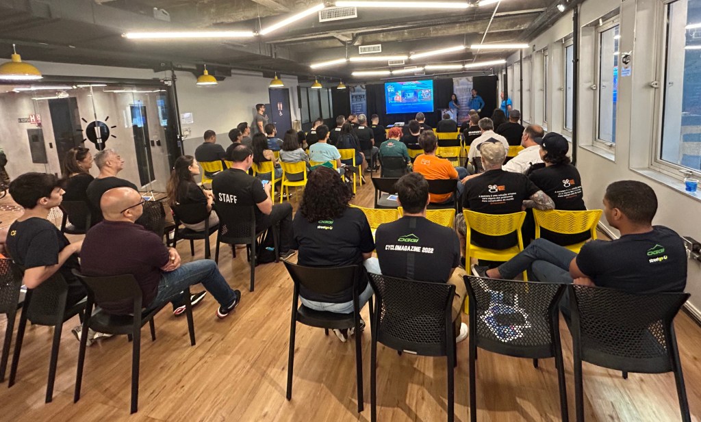 Imagem mostra salão com cadeiras pretas e amarelas ocupadas por pessoas que acompanham apresentação no palco