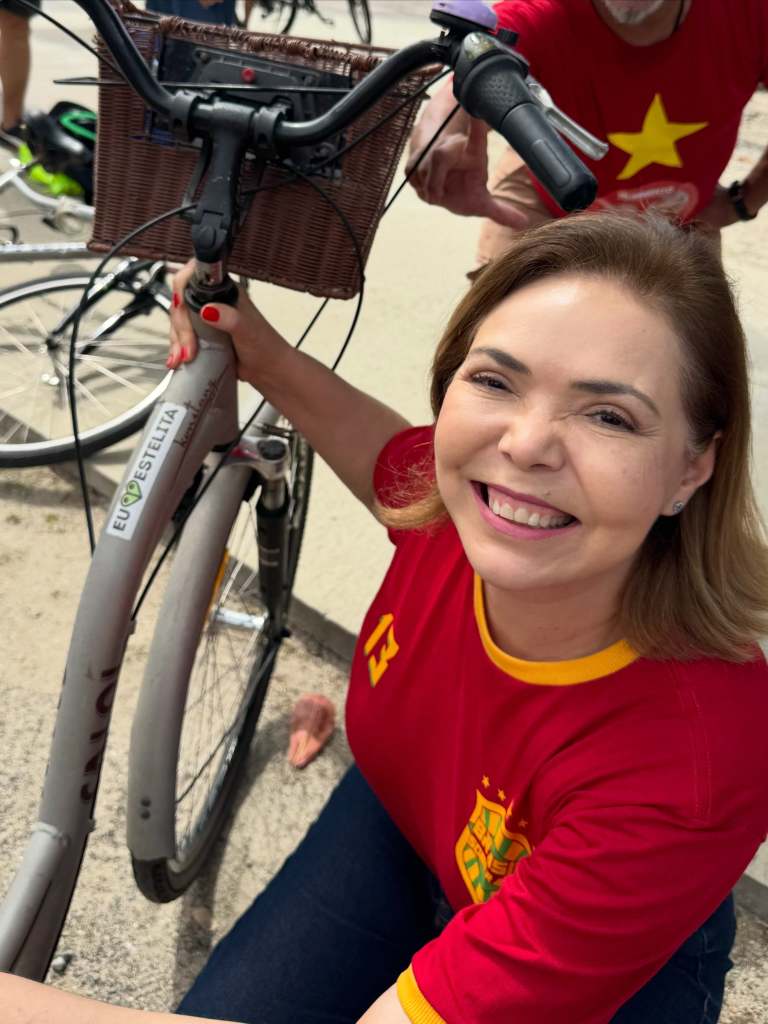 Mulher sorridente com blusa vermelha ao lado de uma bicicleta, segurando o guidão e posando para a câmera.